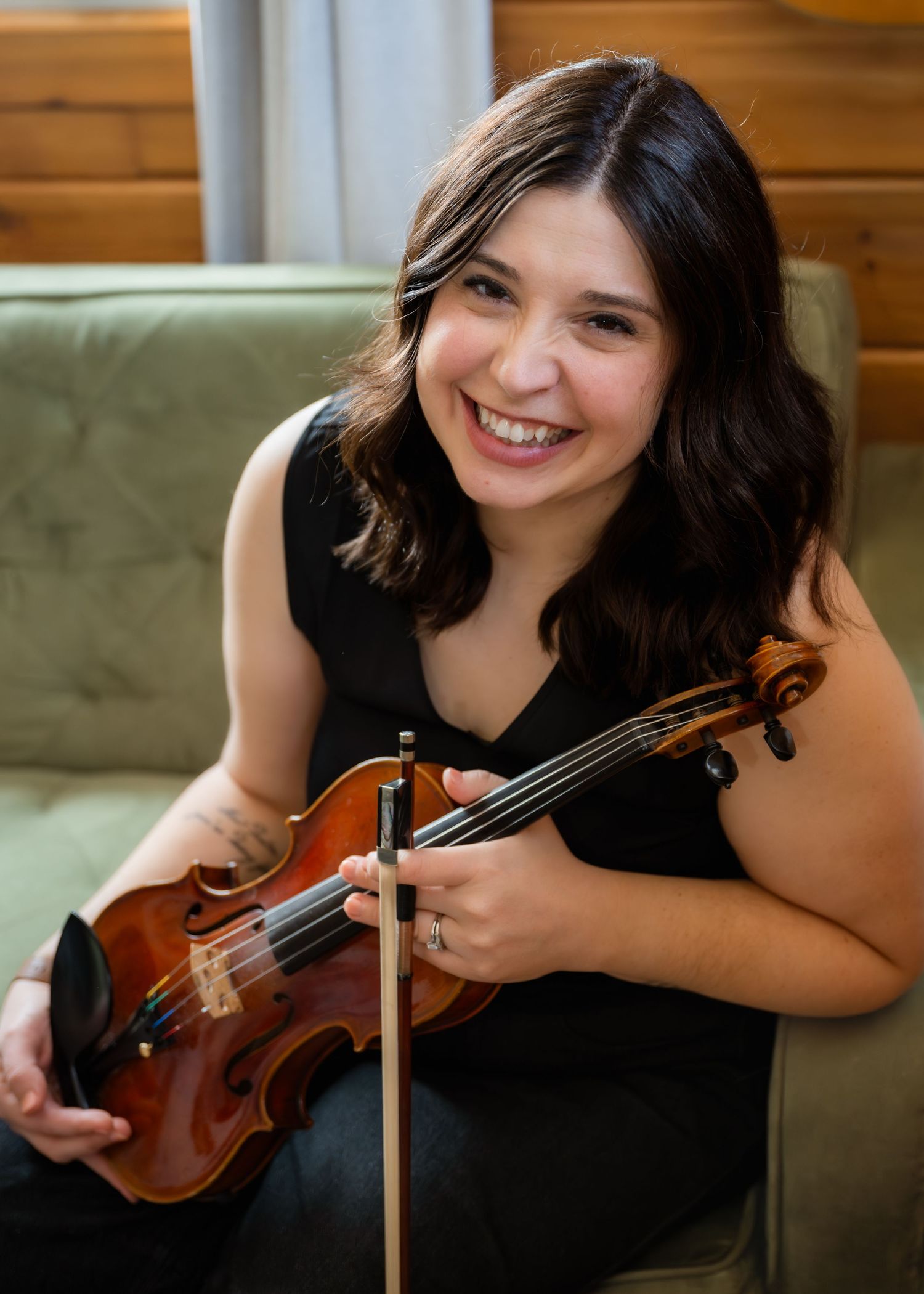 Smiling woman with dark hair poses with a violin and microphone on a wooden table in a cozy studio setting.