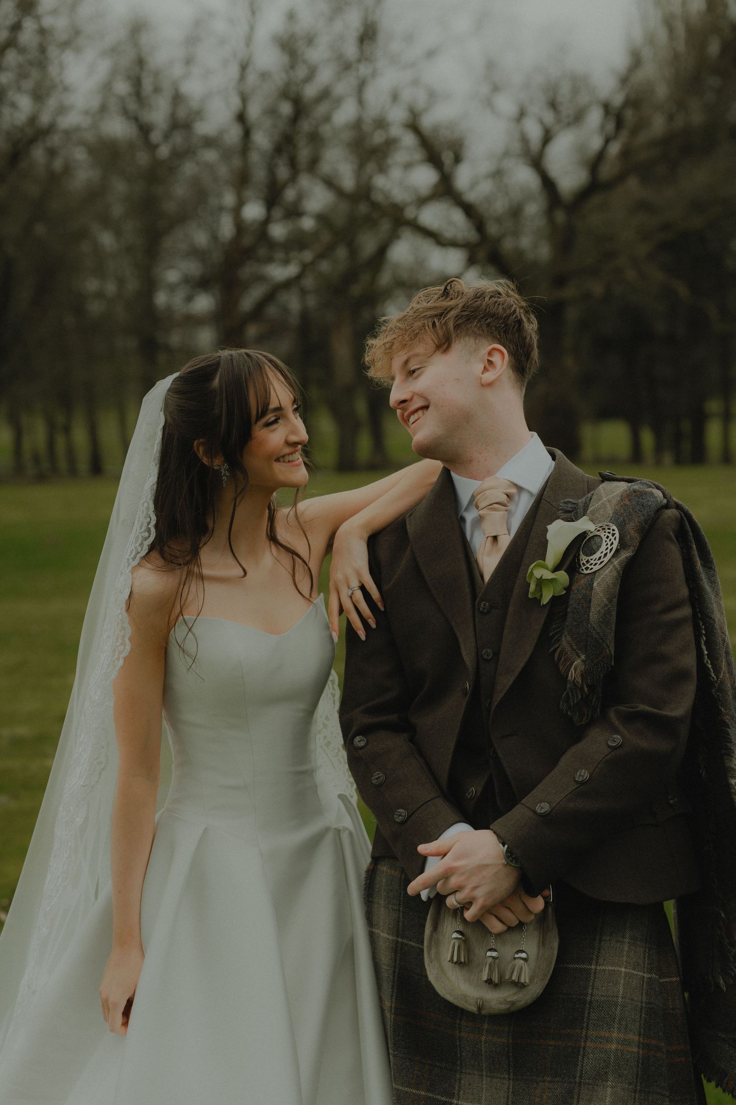 A bride in a white gown and groom in a Scottish kilt share a loving glance outdoors at their wedding.