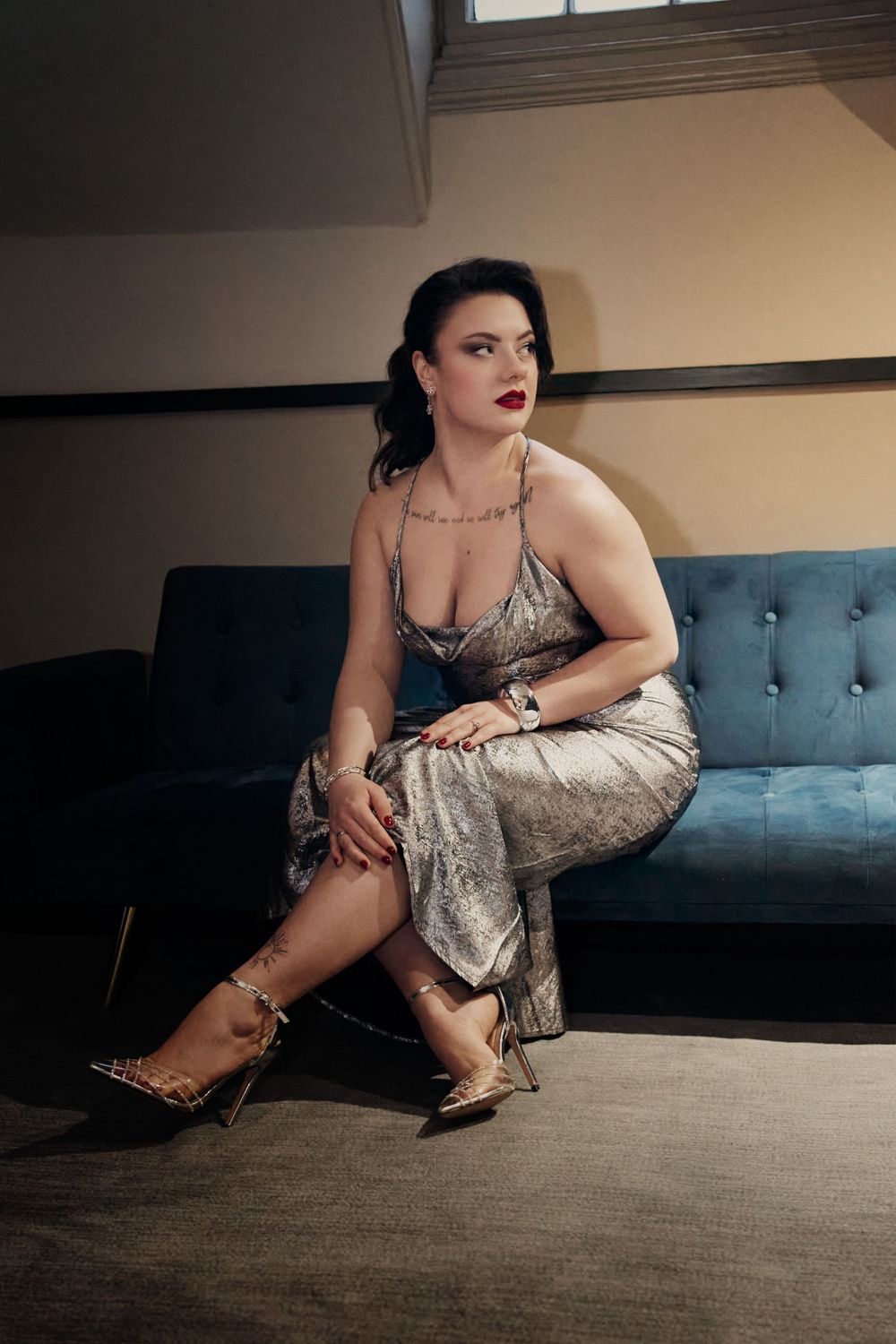 Woman in silver sequined dress seated on blue velvet sofa wearing gold strappy heels.