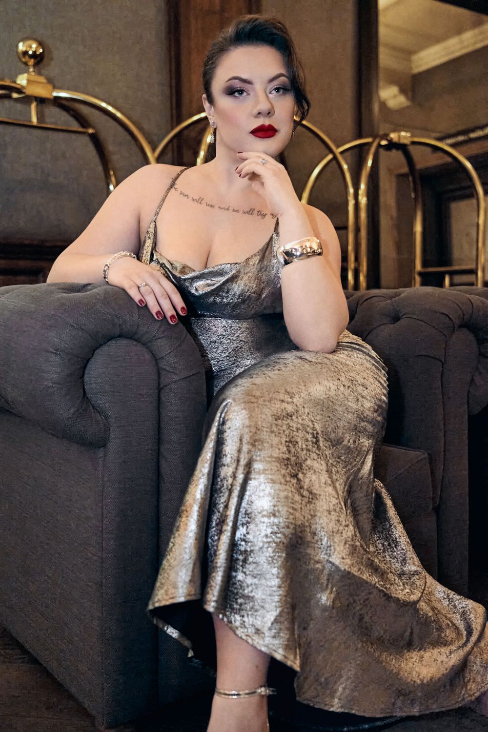 Woman in silver metallic gown seated elegantly on gray tufted chair with gold headboard backdrop.