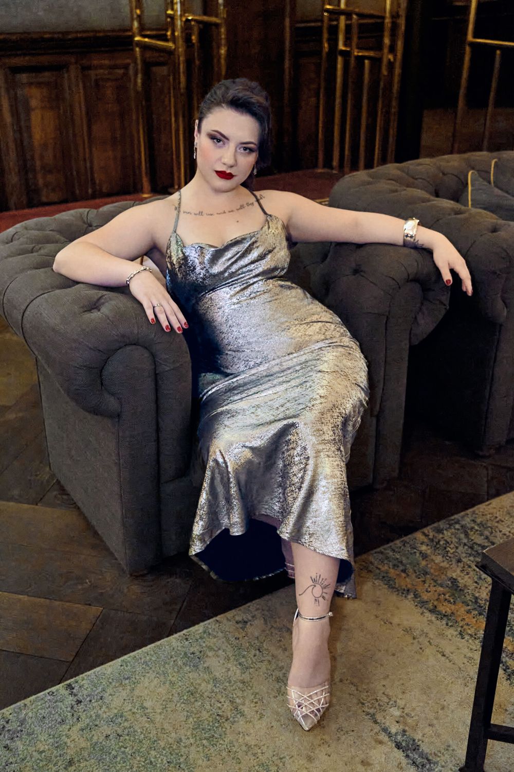 Woman in silver metallic gown seated elegantly in gray tufted armchair, wearing heels and jewelry in upscale interior.