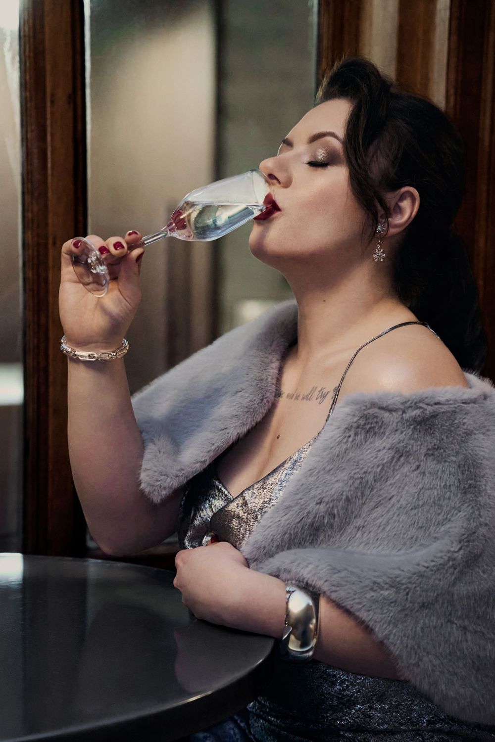 Elegant woman in fur wrap sipping champagne at a table, wearing jewelry and red lipstick in a sophisticated setting.
