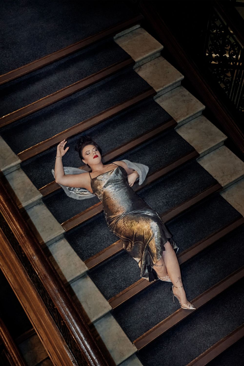 Woman in silver gold gown posed dramatically on grand staircase with dark carpet and ornate wooden railing.
