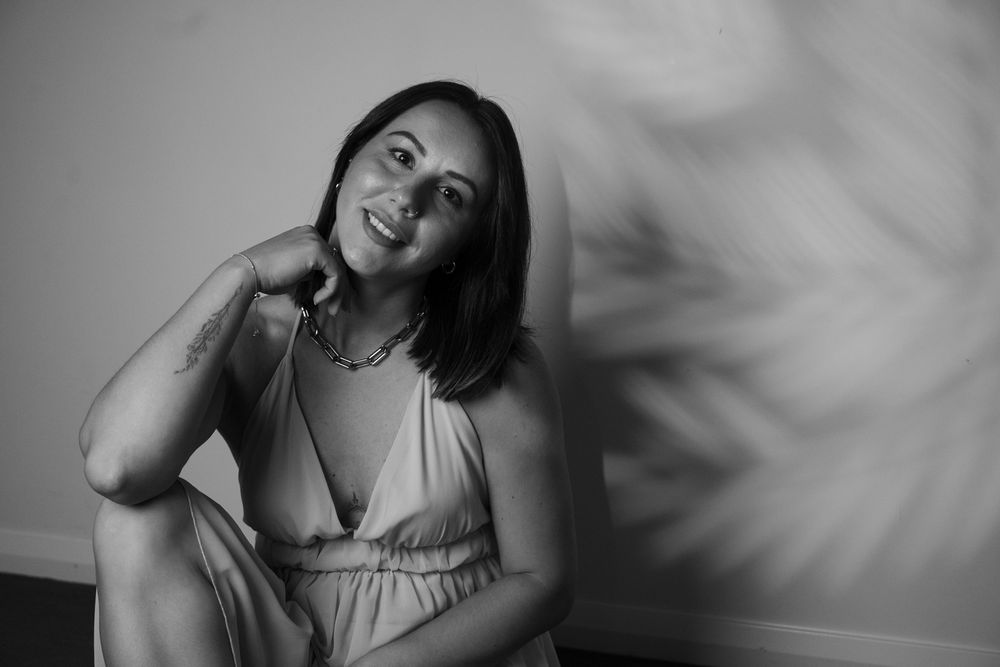 Black and white portrait of smiling brunette woman in spaghetti strap dress leaning against wall.