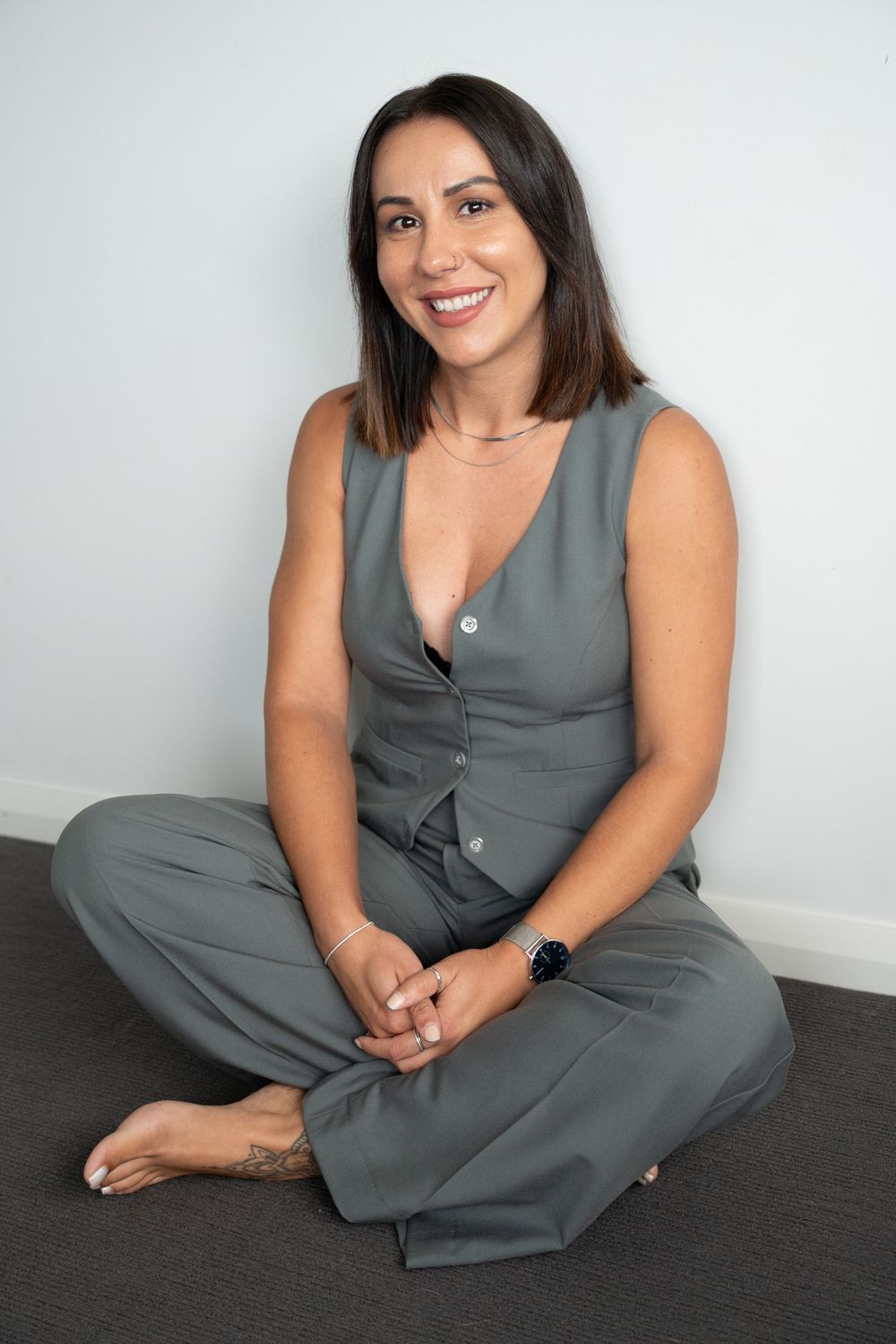 Woman in grey jumpsuit sitting cross-legged on floor smiling at camera in bright studio setting.
