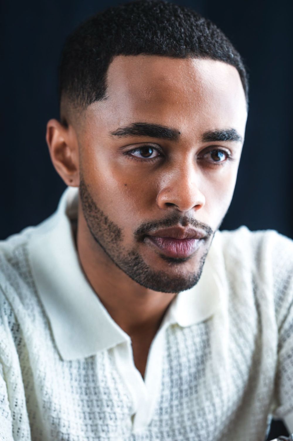 Close-up portrait of a young Black man with a beard wearing a white textured polo shirt.
