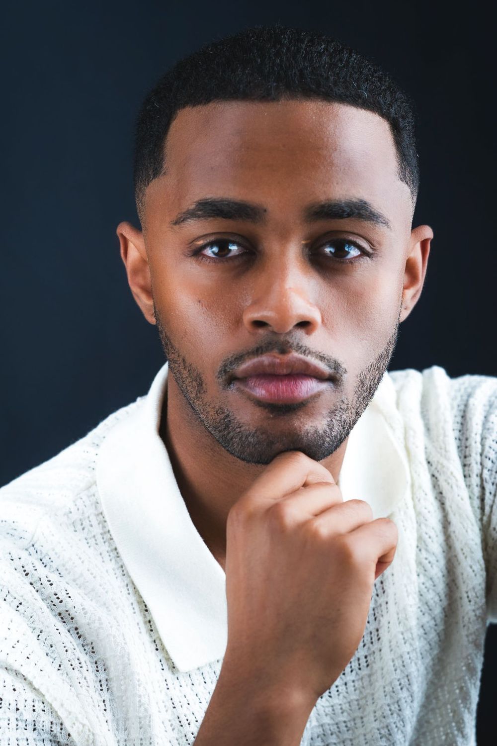 Thoughtful young Black man in a white knit shirt resting chin on hand against dark background.