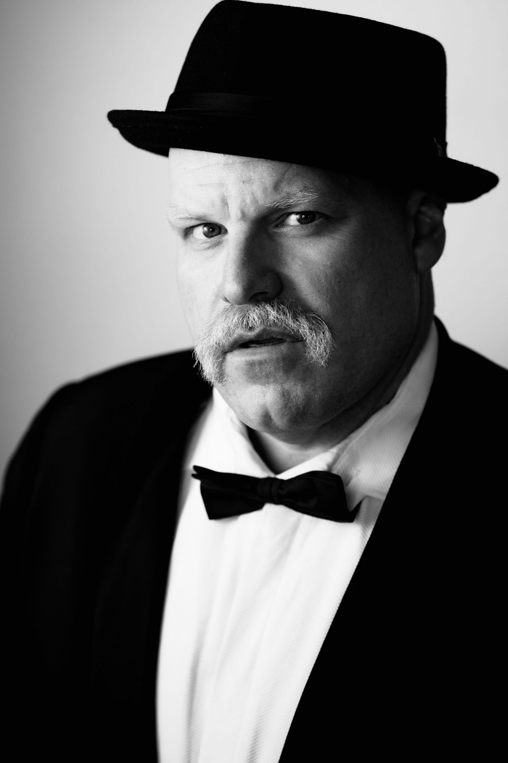 Black and white portrait of mature man with mustache wearing tuxedo, bow tie, and fedora hat.