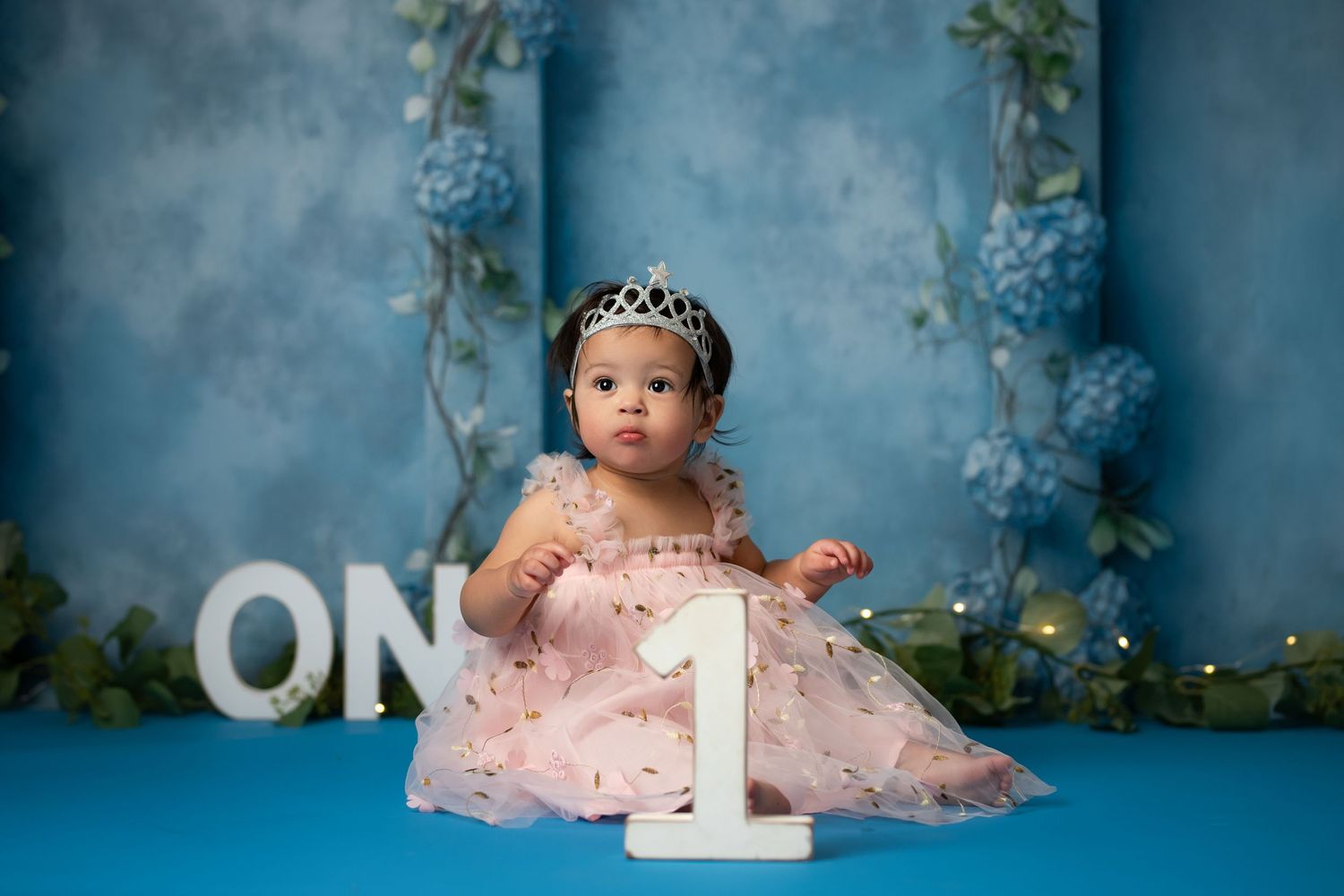 Baby girl in pink tutu dress and tiara sits beside number 1 for first birthday cake smash photo.