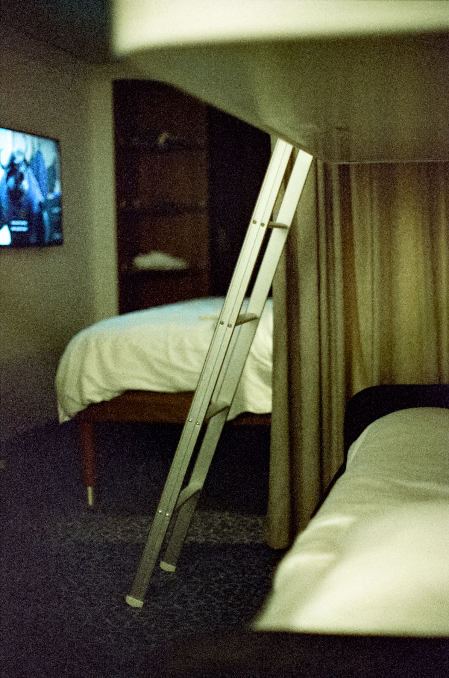 A pair of crutches leaning against a curtain divider between two beds in a dimly lit hotel room with a TV.
