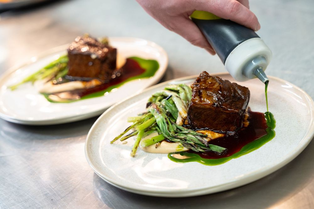 Chef finishing a braised beef short rib dish with green sauce on a white plate in a restaurant kitchen.