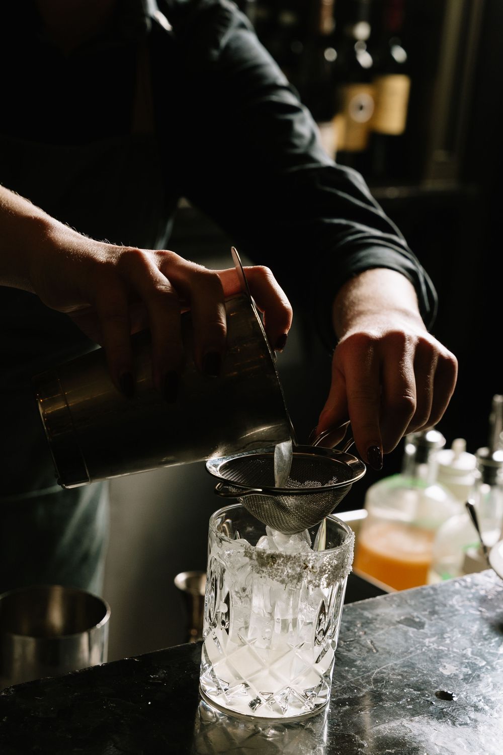 Bartender straining a cocktail through a fine mesh sieve into a crystal glass filled with ice at a dark bar.
