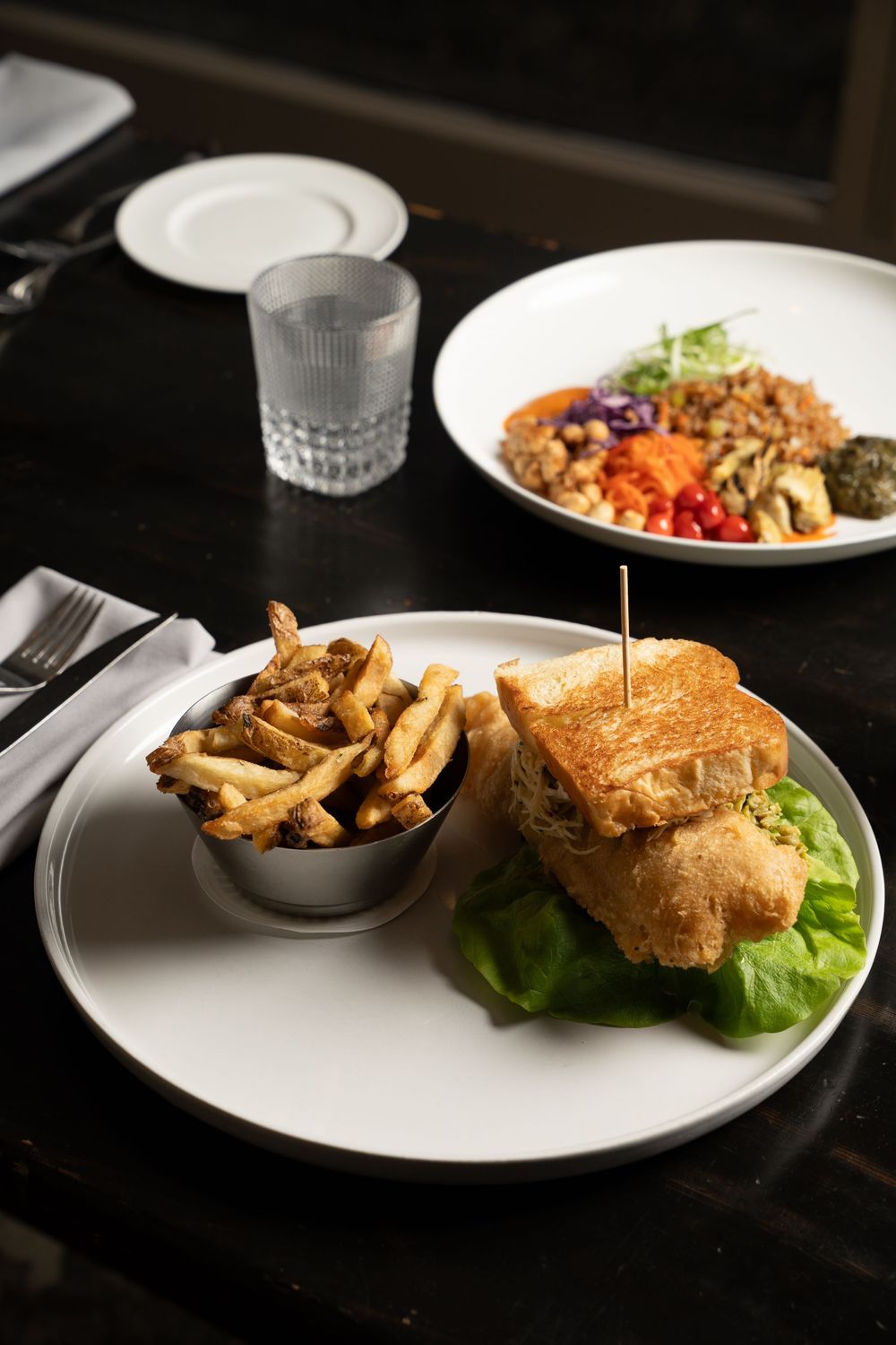 Gourmet sandwich with lettuce on a white plate served with crispy french fries and a grain salad in background.