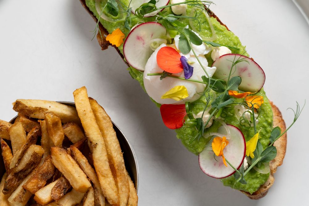 Open-faced sandwich with avocado, radishes, edible flowers, microgreens, served with crispy french fries.