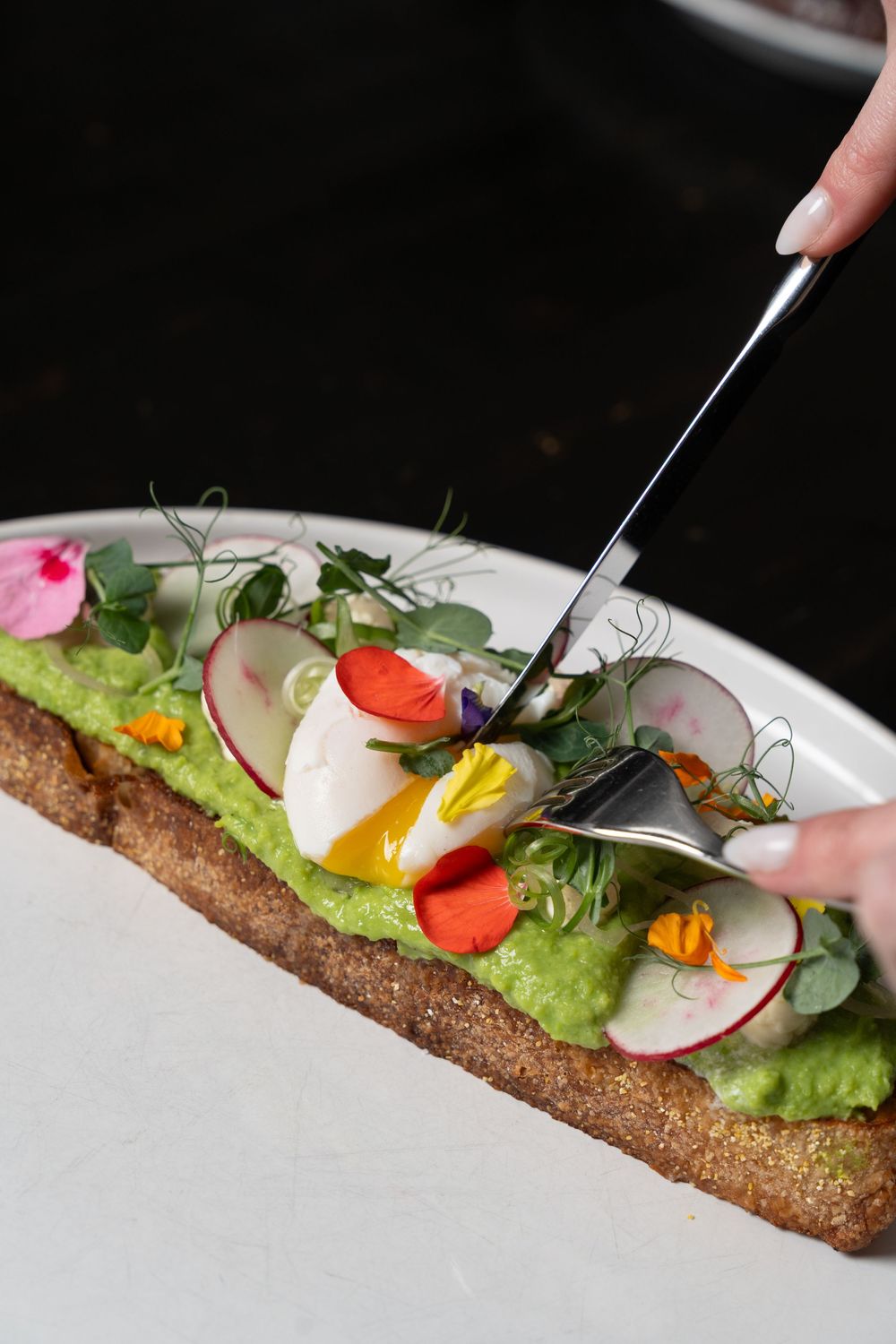 Hand cutting avocado toast topped with radishes, edible flowers, microgreens, and a soft-boiled egg on sourdough bread.
