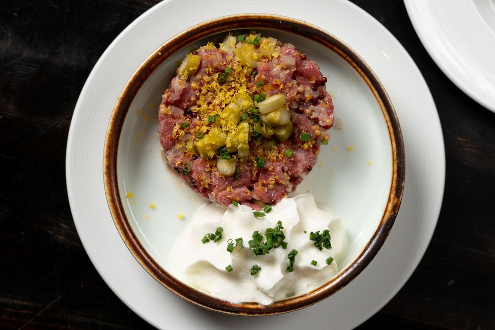 Elegant beef tartare topped with capers and mustard seeds, served with creamy white sauce in a ceramic bowl.