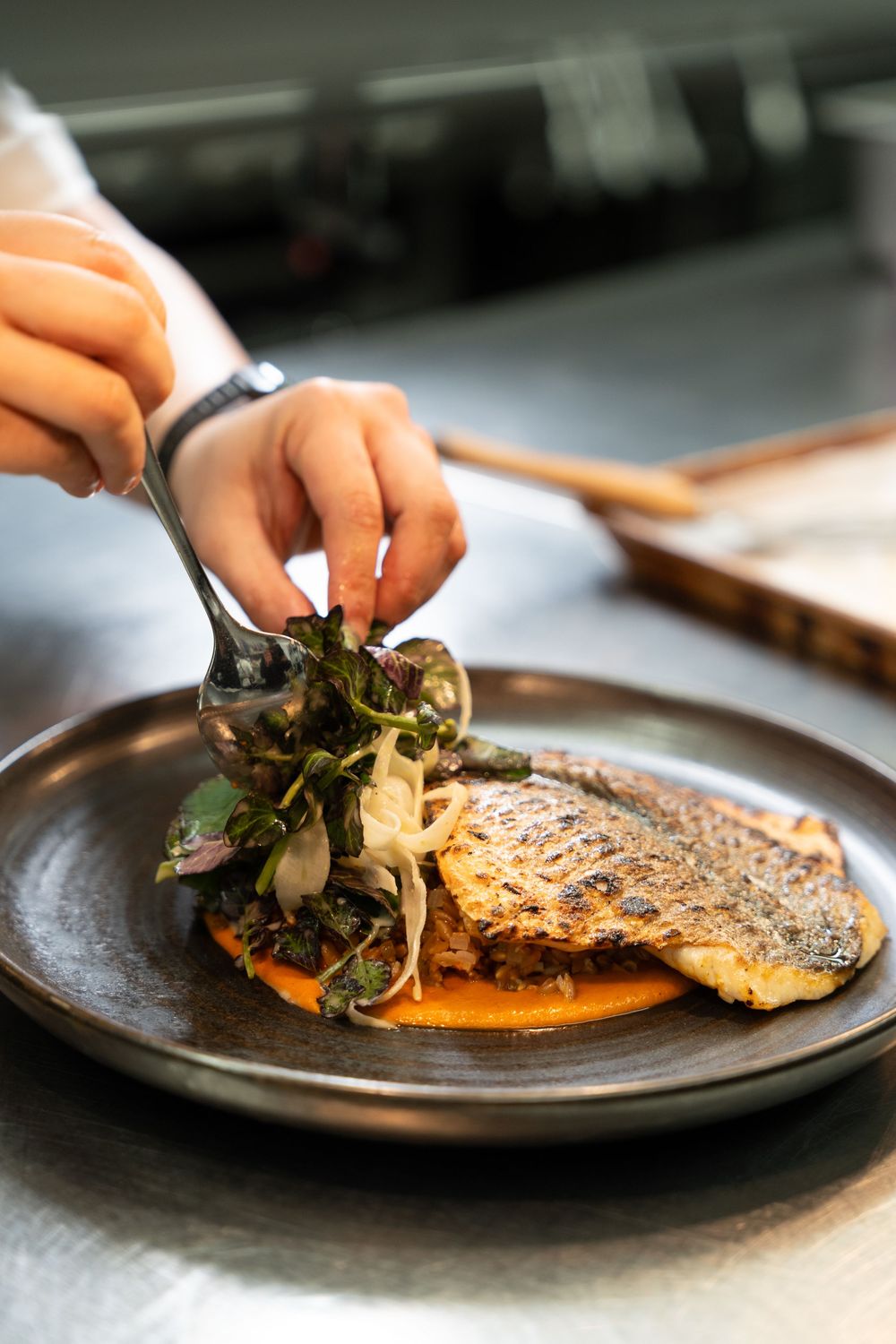Chef plating a crispy pan-seared fish fillet on a dark plate with orange sauce and fresh herb salad.