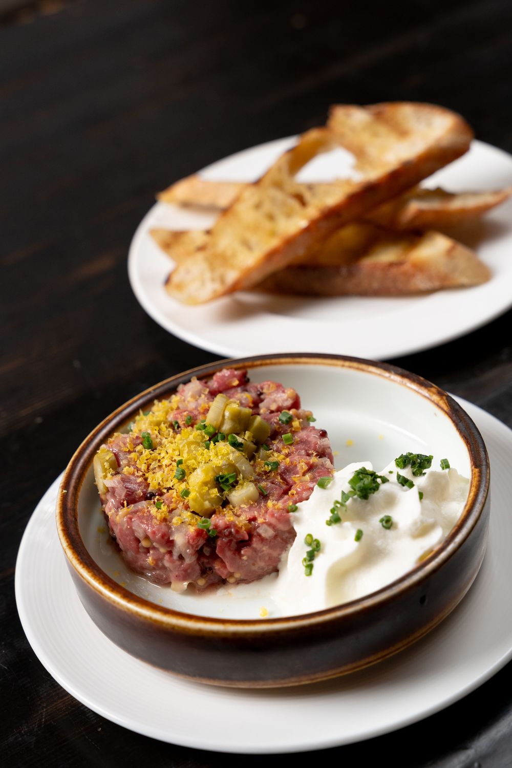 Beef tartare served in a rustic bowl with creamy sauce, capers, and herbs, accompanied by toasted bread slices on a white plate.