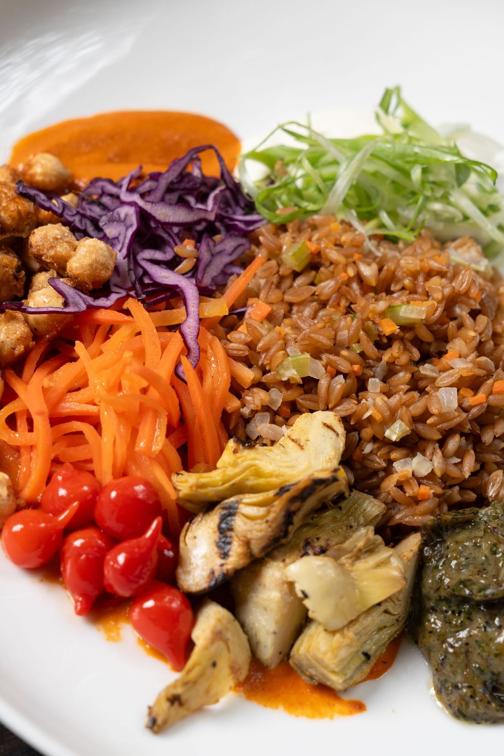 Colorful vegan Buddha bowl with brown rice, shredded purple cabbage, carrots, cherry tomatoes, artichokes, and green salad on white plate.
