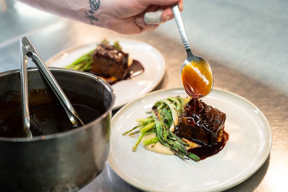 Chef spooning rich brown sauce over braised meat with green vegetables on a white plate in a professional kitchen.