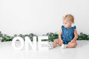 Adorable one-year-old baby girl in denim romper sitting beside large white ONE letters with greenery.