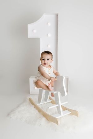 Happy one-year-old baby sitting on white rocking horse in front of large illuminated number one.