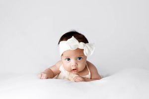 Cute baby girl with white bow headband doing tummy time on white blanket looking at camera.