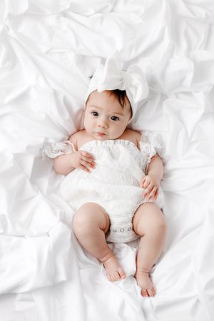 Sweet baby girl in white lace romper and bow headband lying on white wrinkled bedsheet.