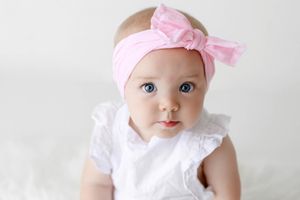 Beautiful blue-eyed baby girl wearing pink bow headband and white ruffled dress portrait.