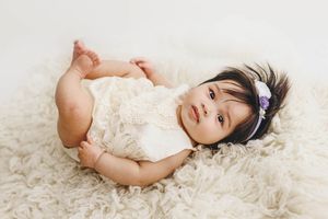 Adorable baby girl in cream lace outfit lying on fluffy rug with purple flower headband.
