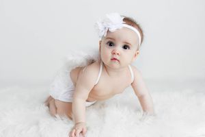 Sweet baby girl in white tutu and floral headband crawling on a soft white fur rug.