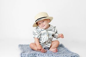 Happy smiling baby boy sitting on a gray knit blanket wearing a straw hat and striped outfit.