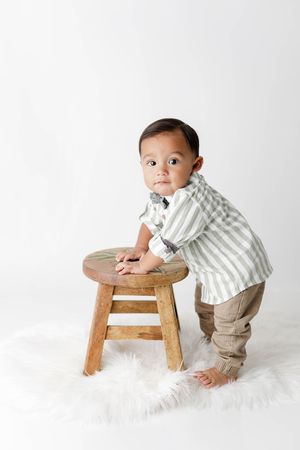 Cute baby boy standing at a wooden stool wearing a striped shirt and tan pants on white fur.