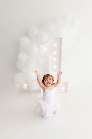 A joyful toddler in a white tutu celebrates her first birthday with arms raised among white balloons and a marquee number one.
