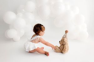 Baby girl in white outfit reaches toward a teddy bear wearing a party hat, surrounded by white balloons.
