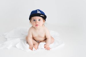 Adorable baby girl wearing an LA Dodgers baseball cap sitting on a white blanket in Menifee photography studio.