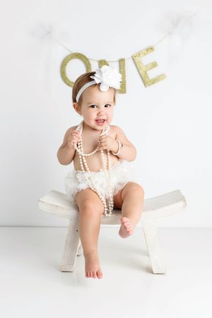 Baby girl in white romper sitting on bench holding pearls with ONE birthday banner.
