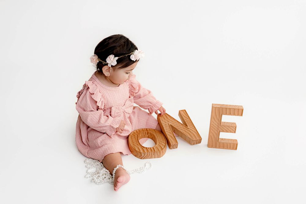 Baby girl in pink dress sitting beside large wooden ONE letters on white background.