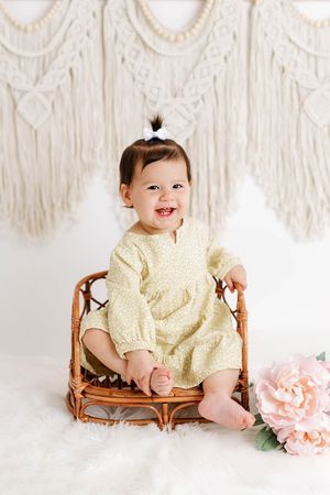 Baby girl's 1st birthday photoshoot in yellow floral dress sitting in rattan chair with macrame backdrop.