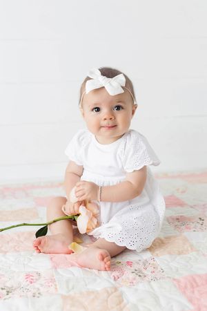 Baby girl in white eyelet dress holding tulip while sitting on floral patchwork quilt.
