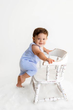 Smiling baby in blue romper standing beside a white vintage bassinet on a white fluffy rug.