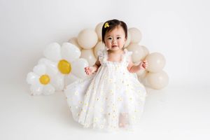 Cute toddler girl in daisy tulle dress surrounded by white and beige balloons.