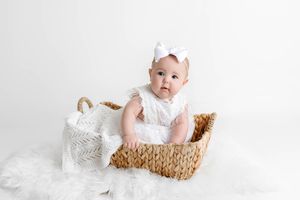 Sweet baby girl in white lace romper sitting inside a woven wicker basket.
