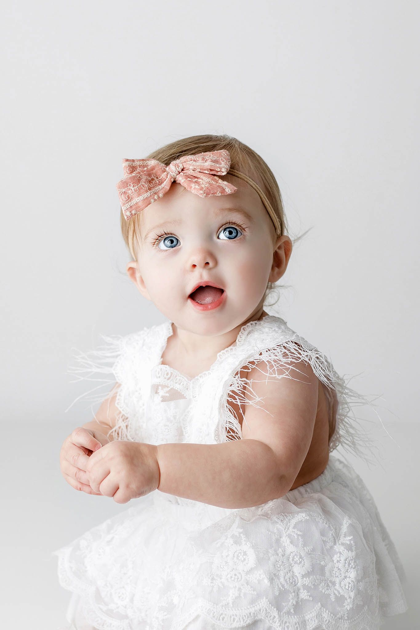 Adorable baby girl in white lace dress with pink bow headband, blue eyes wide open.