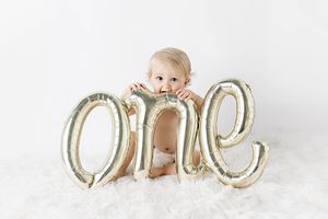 Cute baby sitting behind gold script one balloon on white fluffy rug for 1st birthday photos.