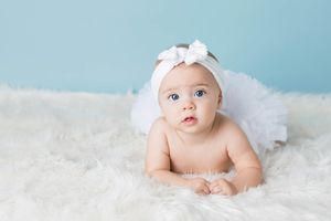 Cute baby girl in white tutu and bow headband doing tummy time on a fluffy white rug.
