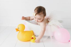 Baby girl in white tutu reaching for a yellow rubber duck beside a pink balloon while sitting in a baby tub.