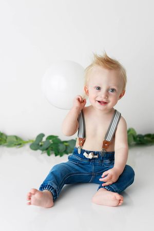 Happy blond toddler boy in jeans and suspenders sitting with a white balloon on white background.