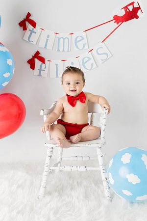 Airplane themed 1st birthday Menifee photoshoot of boy in bow tie sitting on white chair with airplane birthday banner and balloons.