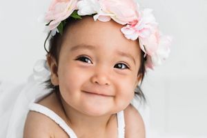 Smiling baby girl wearing a pink floral crown in a bright white studio portrait.